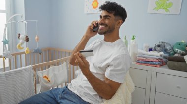 Yatak odasında sakallı genç bir adam bebek beşiğinin yanında kredi kartı tutarken cep telefonu kullanıyor, gülümsüyor ve gelişigüzel giyiniyor..