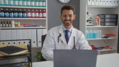 Klinikteki yakışıklı, beyaz önlüklü, steteskopla gülümseyen ve hastane odasında tıbbi malzemeler ve kitaplarla çevrili olan bir adam.