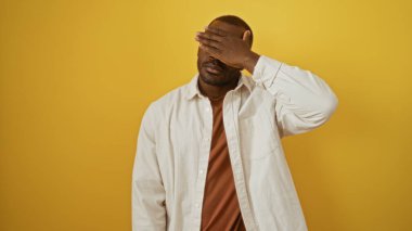 Genç, siyah, bir eliyle yüzünü kapatan adam canlı sarı bir arka planda, beyaz gömlek giyiyor ve duygularını ifade ediyor.