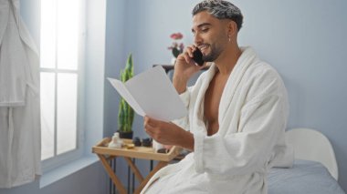 Kaplıca salonunda bornozlu genç bir adam, telefonda konuşurken bir belge okuyor, çevresi huzur ve güzellik öğeleriyle çevrili..