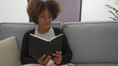 Modern apartman odasındaki rahat koltukta kitap okuyan bir kadın doğal saç stili ve günlük kıyafetlerle rahatlamış bir ortam sergiliyor..