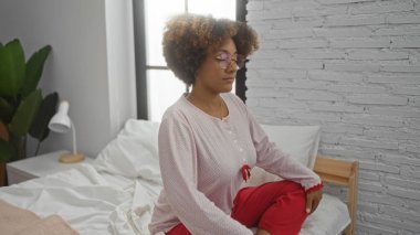 Sakin bir ifadeyle yatakta meditasyon yapan kadın, kıvırcık saçlı ve gözlüklü, günlük kıyafetler giymiş, beyaz tuğlalı duvarlar ve doğal ışık içeren parlak bir yatak odasında..