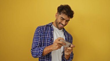 Akıllı telefonuyla oynayan sakallı genç adam canlı sarı bir arka planda gülümsüyor ve odaklanmış görünüyor, gelişigüzel bir ortamda eğleniyor..