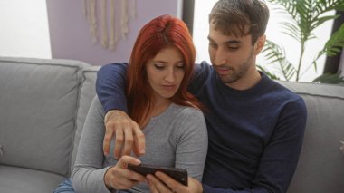 Oturma odasında kanepede oturan bir çift, kadına akıllı telefon gösteren bir adam, sıcak bir evin içinde samimi bir anı yakalıyorlar..