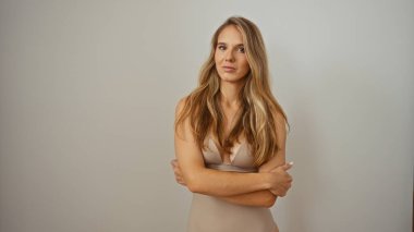 Beyaz bir duvarın karşısında duran çekici sarışın kadın, kendinden emin ve sakin görünüyor, kolları göğsünde çaprazlanmış açık elbiseler giyiyor.