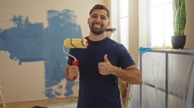 Gülümseyen genç adam, evin yeni boyanmış oturma odasında boya silindiri tutarken, onarım ya da başarı öneriyor.