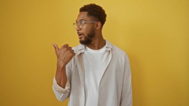 Genç adam sağını işaret ediyor, gözlüklü ve beyaz gömlekli, izole edilmiş sarı arka plan duvarının üzerinde duruyor, ağzı açık şaşırmış görünüyor.