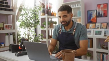 Ev dekorasyonları ve iç mekan bitkileriyle dolu bir mağazada dizüstü bilgisayarda çalışan yakışıklı genç adam çağdaş ve yaratıcı tasarım unsurlarını sergiliyor.