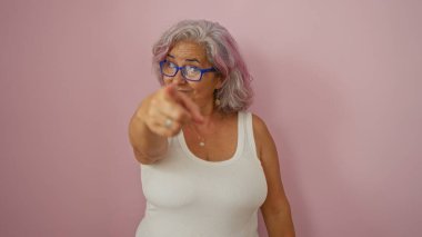 Gri saçlı ve gözlüklü orta yaşlı bir kadın pembe arka planda izole edilmiş kamerayı işaret ediyor.