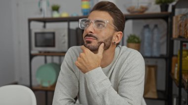 Ofis ortamında düşünen, gözlük takan, modern dekorlar ve mutfak malzemeleriyle çevrili, düşünceli bir yüz ifadesi olan genç bir adam..