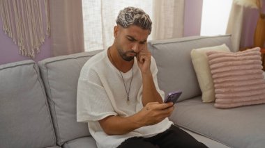 Evdeki oturma odasında sakallı genç bir adam, beyaz bir gömlek giyerken düşünceli bir ifadeyle akıllı telefona bakıyor.