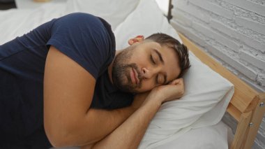 Latin kökenli genç adam rahat yatak odasında beyaz yastıkta huzur içinde uyuyor. Doğal ışık altında rahat ifadesini ve rahat ev atmosferini vurguluyor..