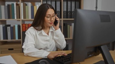 Ofis ortamında bilgisayar kullanırken telefonla konuşan genç bir kadın. Etrafı kitaplık ve dosya klasörleriyle çevrili. Mesleki faaliyeti gösteren kapalı bir çalışma ortamı.