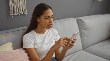 Evinin oturma odasında akıllı telefon kullanan genç bir kadın.