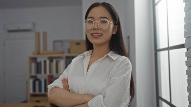 Beyaz tişörtlü genç Çinli kadın modern ofiste kollarını kavuşturmuş duruyor..
