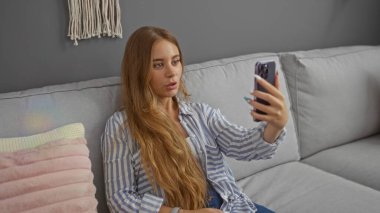 Uzun sarı saçlı, genç ve çekici bir kadın modern bir oturma odasında selfie çekiyor.