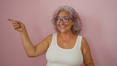 Gülümseyen ve gözlük takan bir kadın pembe arka plana işaret ederken gri saçlarını ve canlı kişiliğini sergiliyor.