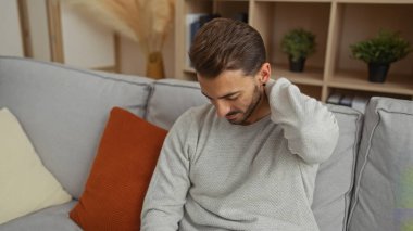 Evdeki genç adam, modern bir oturma odasındaki kanepede oturan boynu ağrıyan rahat ama acılı bir ortamda rahatlamış bir ifadeyi yansıtıyor..