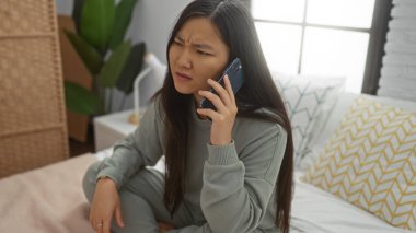 Kadın rahat bir yatak odasında, yumuşak yastıklar ve bitkilerle, rahat ve rahat bir atmosferi taşırken akıllı telefonuyla konuşuyor..