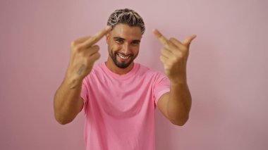 Sakallı, pembe gömlekli, pembe arka planda duran, saldırgan bir el hareketi yapan genç, gülümseyen Arap adam.