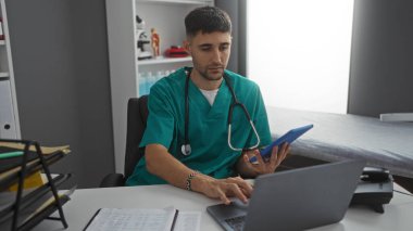 Steteskoplu önlük giyen genç adam modern klinikte dizüstü bilgisayarla çalışıyor.