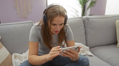 Kulaklıklı kadın rahat oturma odasında akıllı telefona odaklanmış, günlük kıyafetler giymiş, modern koltuğa oturmuş, yumuşak yastıklarla ve ev bitkileriyle çevrili..