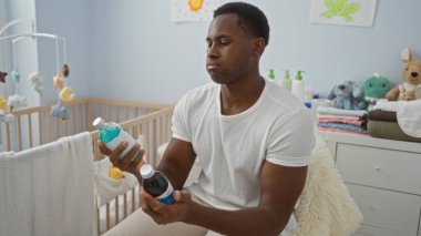 Yatak odasında oturan genç adam elinde şişelerle, arka planda bebek beşiği, evde düşünceli ve ilgili bir ruh halini yansıtıyor..