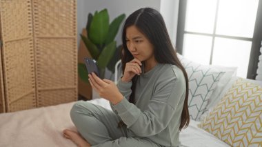 Yatak odasında, akıllı telefon kullanan, rahat kıyafetler giyen, yastıklarla çevrili ve hafif ışıklandırmayla çevrili bir kadın..