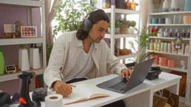 Uzun saçlı yakışıklı İspanyol bir adam dizüstü bilgisayarda çalışıyor ve çeşitli dekorasyon malzemeleriyle dolu bir dekor dükkanında deftere yazı yazıyor.