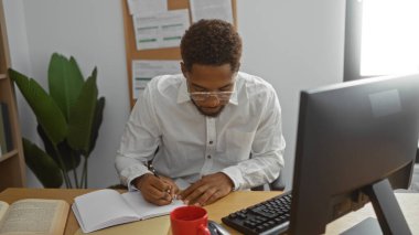 Ofisteki masada bilgisayar, kırmızı kahve fincanı ve arka planda görünen bitkilerle yazan genç adam.