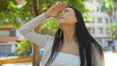 Şehir parkında güneş ışığından gözlerini gölgeleyen bir kadın güneşli bir günde şehir ortamının tadını çıkaran genç ve çekici bir Çinli kadını canlandırıyor..