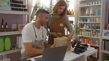 Çalışanlar ev dekorasyon mağazasında cep telefonlu paketleri tarıyor, adam masada oturuyor, kadın yardımcı oluyor, dekoratif eşyalarla çevrili, raflar ev dekorasyonunda ürünlerle dolu.