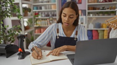 Ev dekorasyonu mağazasında çalışan genç bir kadın masanın üzerinde dizüstü bilgisayar ve barkod tarayıcı ile notlar alıyor..