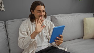 Uzun saçlı, gülümseyen ve tabletin ekranına el sallayan genç bir adam bir video görüşmesi sırasında oturma odasında kanepede oturuyor.