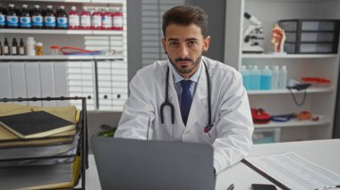 Beyaz önlüklü ve steteskoplu bir hastane ofisinde oturan doktor arka planda tıbbi malzemeler ve organize dosyalar var..