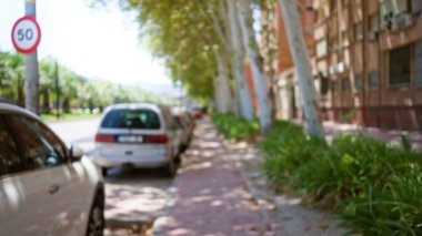 Bulanık bir sokak sahnesi. Bulanık bir kaldırım boyunca çözünmüş arabalar. Güneşli bir günde arka plan efekti olan büyük yapraklı ağaçlar tarafından gölgeleniyor.