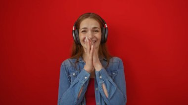 Canlı kırmızı bir duvara karşı kulaklıkla gülümseyen bir kadın, izole, renkli bir ortamda neşeli bir an sergiliyor, mutluluğu ve modern tarzı ifade ediyor..