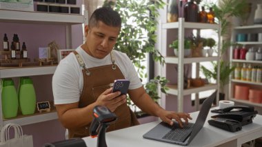 Fırında çalışan genç adam dizüstü bilgisayar ve telefon kullanarak raflarda çeşitli ürünler kullanıyor.