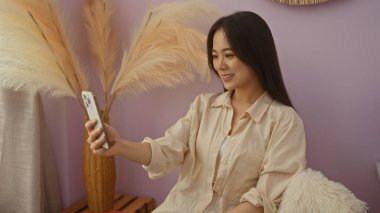 Yumuşak ışıklandırması ve iç dekorasyonu olan rahat bir oturma odasında akıllı telefon kullanarak gülümseyen Çinli genç bir kadın.