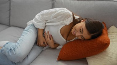 Oturma odasında rahatsız bir şekilde karnını tutarak kanepede yatan genç bir kadın.