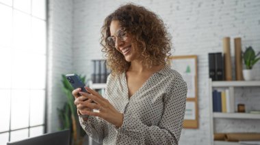 Ofiste gülümseyen, gözlüklü, akıllı telefonu geniş pencerenin yanında tutan, rafları ve bitkileri olan, profesyonel ama rahat bir ortam sergileyen bir kadın..