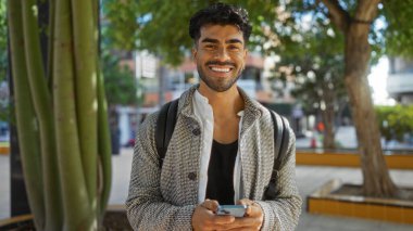 Şehir parkında telefon ve sırt çantasıyla gülümseyen yakışıklı İspanyol adam ağaçlar ve modern binalarla çevrili güneşli bir günün tadını çıkarıyor..