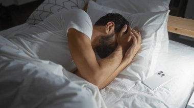 Yatağında akıllı bir telefonla yatak odasında yatan sakallı bir İspanyol genç adam.