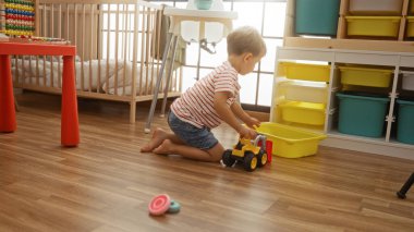 Parlak ışıklandırılmış bir oyun alanının zemininde oyuncak bir kamyonla oynayan küçük çocuk, renkli depolama kutuları ve beşikle çevrili, çizgili gömlek ve kot şort giyiyor.