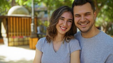 Parkta gülümseyen bir çift, arka planda şehir ortamı olan ağaçların altında sevgi ve ilişki sergiliyor.