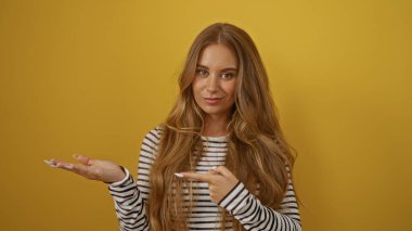 Güzel sarışın kadın, izole edilmiş sarı arka plan duvarına gülümseyip kameraya bakan çizgili bir gömlek giyerek el hareketleriyle işaret ediyor.