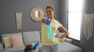 Oturma odasını sprey şişeyle temizleyen genç bir kadın. Parlak apartman dairesinde güvenle gülümsüyor.