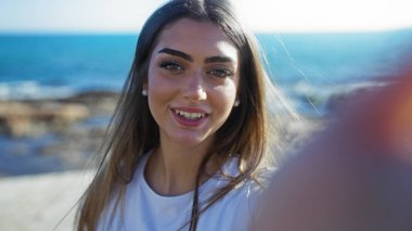 Güzel, genç, İspanyol bir kadın güneşli bir günde deniz kenarında arka planda okyanusla birlikte selfie çekiyor..