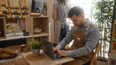 İçerde dizüstü bilgisayarda çalışan bir adam ve fırın arka planında önlüklü bir kadın.