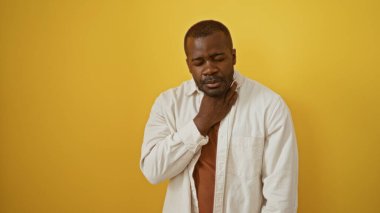 İzole edilmiş sarı bir arka planı olan yakışıklı genç bir adam boğazı ağrıyor ve günlük kıyafetler giyiyor.